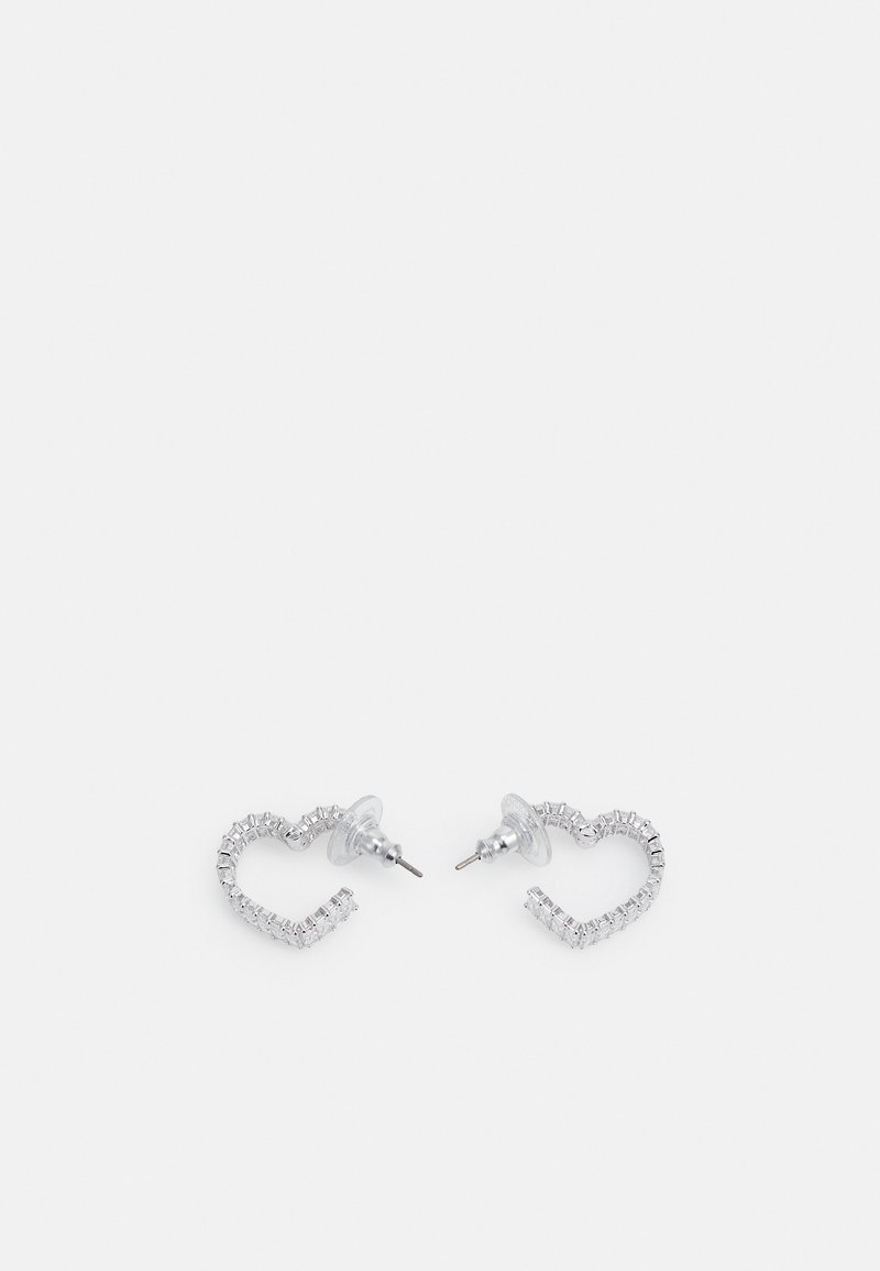 Boucles d'oreilles clous en forme de cœur ton argent avec cristaux clairs taillés en carré et attaches plastiques transparentes sur fond blanc.