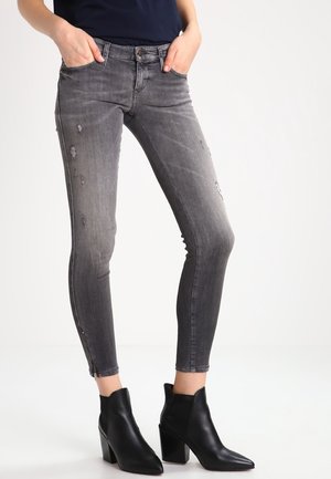 Schmal geschnittene, ausgebleichte graue Jeans mit Used-Details, Vorder- und Gesäßtaschen, Reißverschlüssen am Knöchel, getragen mit schwarzen Stiefeletten mit Absatz.