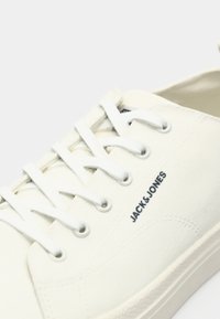 Bílé plátěné tenisky se zaoblenou špičkou, texturovaným povrchem a plochými bílými tkaničkami. Obsahují šité prvky a malé černé logo značky.