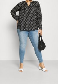 Sort langærmet bluse med hvidt geometrisk mønster, parret med lyseblå tætsiddende jeans og hvide blokhæle sandaler, mens der holdes en sort taske.