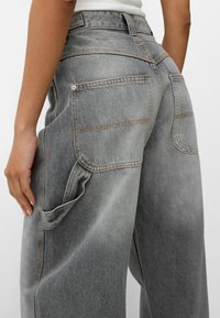 Grå denim bredbenta jeans med synlig sömnad, två bakfickor och en sidoficka. Tyget har en mjuk, lätt sliten textur.
