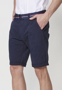 Man in een marineblauwe opgerolde short met een riem met rood, wit en blauwe strepen, met een hand in zijn zak, staand tegen een witte achtergrond.