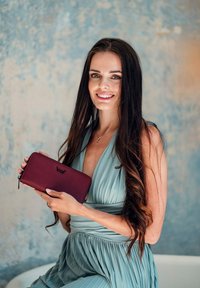 Burgundská kožená peněženka s texturovaným povrchem, zipovým uzávěrem a logem. Modelka má na sobě jemné modré skladané šaty na texturovaném pozadí.