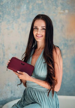 Burgundská kožená peněženka s texturovaným povrchem, zipovým uzávěrem a logem. Modelka má na sobě jemné modré skladané šaty na texturovaném pozadí.