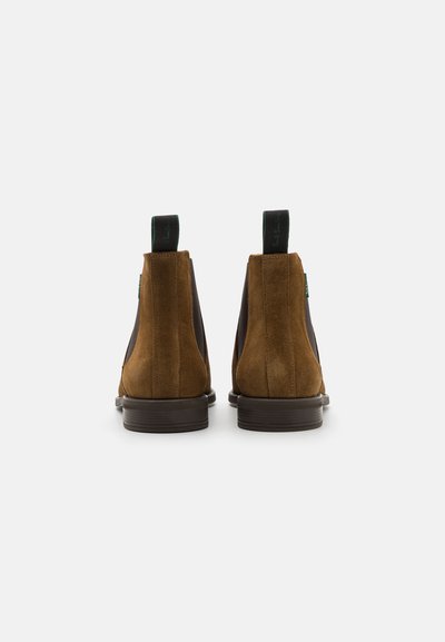 Bottes chevaucheuses en daim marron avec des panneaux latéraux en élastique, une semelle lisse foncée et des tirettes ornées d'accents noirs et verts.