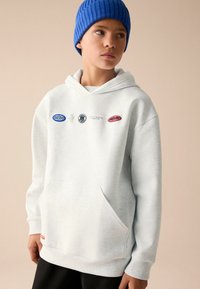 Haut à capuche gris clair avec poche avant, arborant des logos colorés sur la poitrine. Accompagné d'un bonnet en tricot côtelé bleu. Texture lisse.