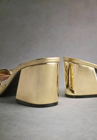 Auksinės platforminės bateliai su blokiniais kulnais, turintys lygų metalinį paviršių ir apvalias briaunas. Padas juodas, sukuriantis kontrastą su auksine spalva.