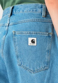 Lys blå denim jeans med en synlig baklomme som har en hvit Carhartt-lapp. Tøyet har en teksturert overflate og kontraststing.