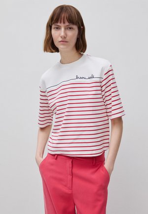 Femme aux cheveux bruns courts portant un t-shirt blanc à rayures rouges avec le texte « bisou salé » et un pantalon rose vif, mains dans les poches, fond neutre.