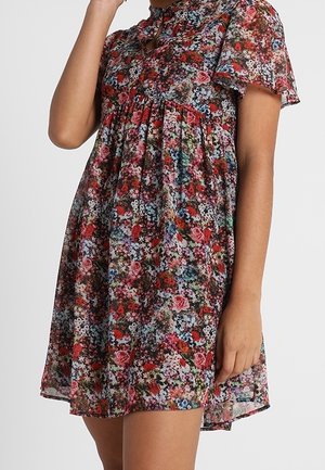 Femme portant une robe courte et ample à imprimé floral avec des manches bouffantes et une taille froncée, debout devant un fond clair uni.