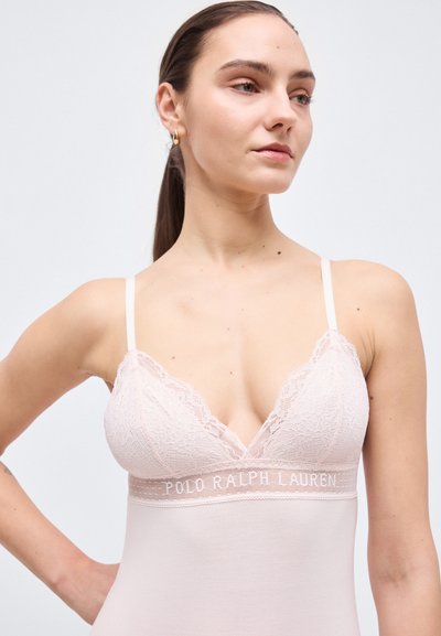 Γυναίκα με σκούρα μαλλιά σε κοτσίδα που φοράει ένα ανοιχτό ροζ δαντελένιο τοπάκι με το κείμενο "Polo Ralph Lauren" στη ζώνη στο στήθος.