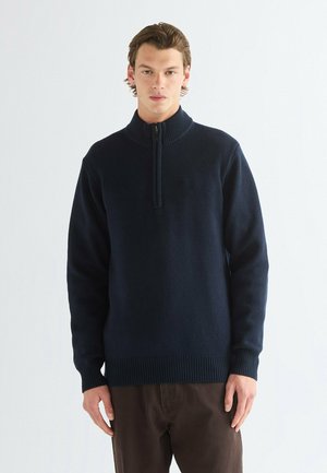 Jeune homme portant un pull bleu marine à demi-fermeture éclair et un pantalon marron, debout devant un fond clair uni, faisant face à l’avant.