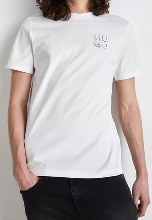 Personne portant un T-shirt blanc uni à manches courtes avec le logo "HUGO" sur la poitrine, associé à un pantalon noir, debout avec une main dans la poche.