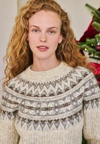 Kötött pulóver, amelynek krém alapja van, barna és szürke geometrikus mintákkal. Vastag, textúrált fonal és kerek nyakkivágás.