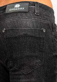 Jeans in denim nero con un primo piano di una tasca posteriore, dettaglio di rivetti argentati e un'etichetta con marchio in vita. Texture liscia, design aderente.