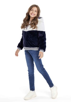 Ragazza giovane che sorride, indossando una giacca in pelliccia sintetica blu navy e bianca con cappuccio, jeans blu e sneakers bianche, in piedi con un piede leggermente sollevato.