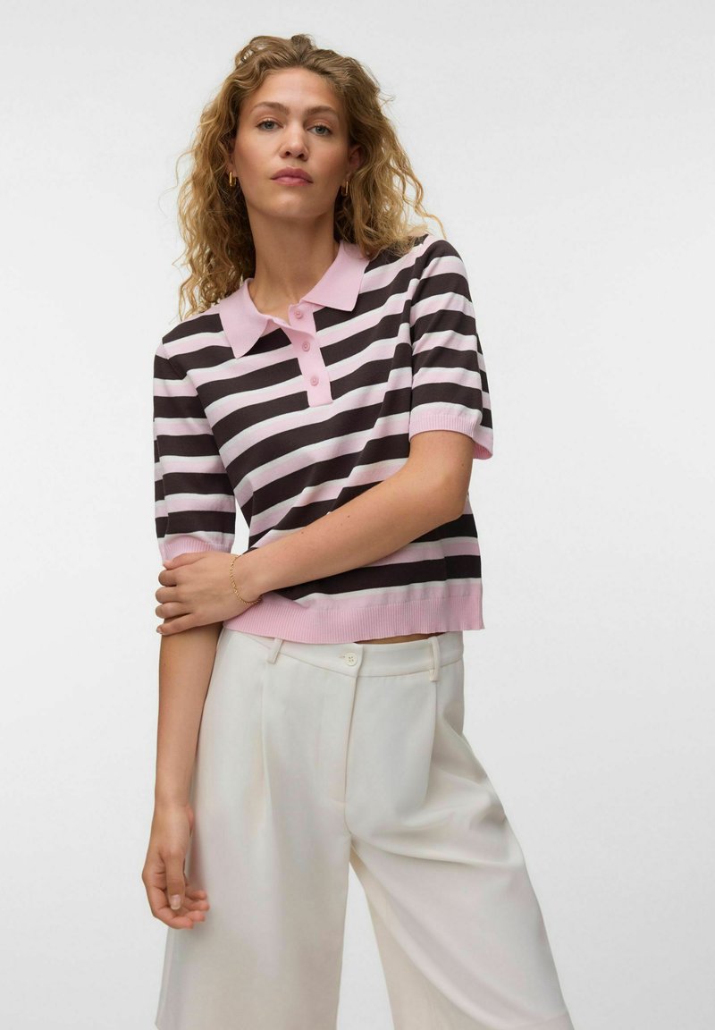 Femme aux cheveux blonds bouclés portant un polo à manches courtes rayé rose et noir et un pantalon blanc sur un fond uni.