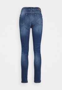 Jeans ajustados em denim azul escuro. Apresenta uma textura suave, cinco bolsos e costura visível. Detalhes desbotados ao longo das pernas e da bainha.