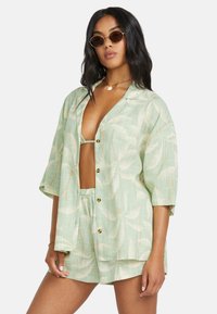Camicia verde chiaro con bottoni e pantaloni corti abbinati con un motivo di foglie di palma. Maniche corte, vestibilità rilassata e bottoni dorati.