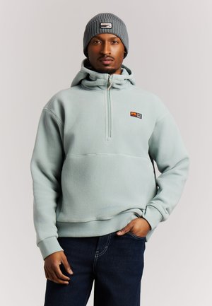 Man die een lichtblauwe fleece hoodie met halve ritssluiting en een grijze gebreide muts draagt, staat met één hand in de zak tegen een effen achtergrond.