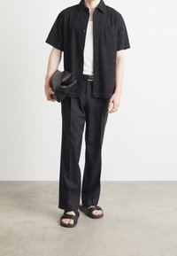 Zwarte shirt met korte mouwen van textiel, witte tanktop eronder, zwarte broek en zwarte sandalen met banden. Houdt een zwarte tas vast.