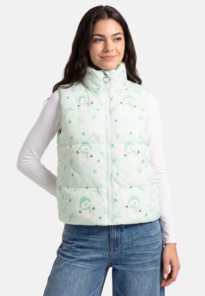 Giovane donna che indossa un gilet imbottito verde chiaro con motivi di pupazzi di neve e fiocchi sopra una camicia bianca a maniche lunghe e jeans blu.