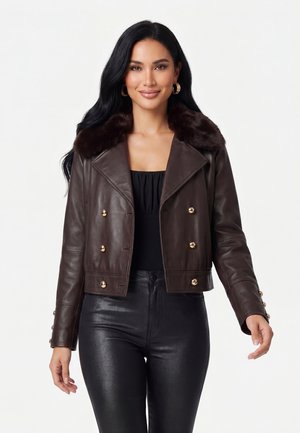 Vrouw met lang zwart haar die een donkerbruine leren jas met een bontkraag en gouden knopen draagt, gecombineerd met een zwarte broek en oorringen.