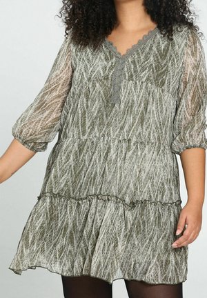 Kobieta ubrana w zieloną, wzorzystą, warstwową sukienkę z półprzezroczystej tkaniny, z rękawami 3/4 zakończonymi bufkami i dekoltem w kształcie litery V wykończonym koronką, stojąca na jednolitym tle.