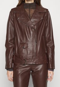 Donna con una giacca in pelle marrone scuro da biker con dettagli in zip e pantaloni in pelle abbinati, sopra una maglietta nera trasparente.