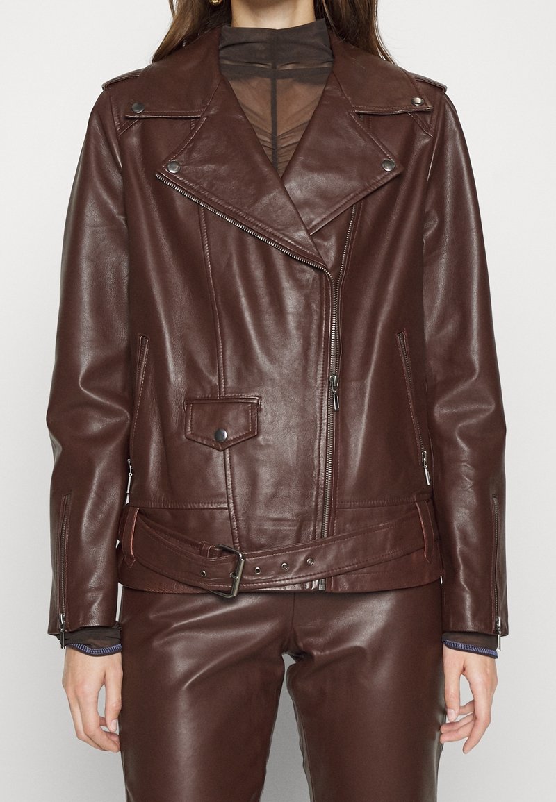Donna con una giacca in pelle marrone scuro da biker con dettagli in zip e pantaloni in pelle abbinati, sopra una maglietta nera trasparente.