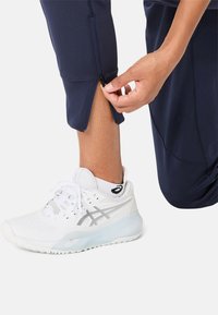 Marineblauwe sportbroek met ritssluitingen aan de boorden, gecombineerd met witte hardloopschoenen met reflecterende accenten en textuur in het bovenwerk van mesh.
