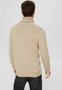 Pull en maille beige avec col montant, présentant une surface texturée et des manches ajustées, associé à un jean en denim foncé.