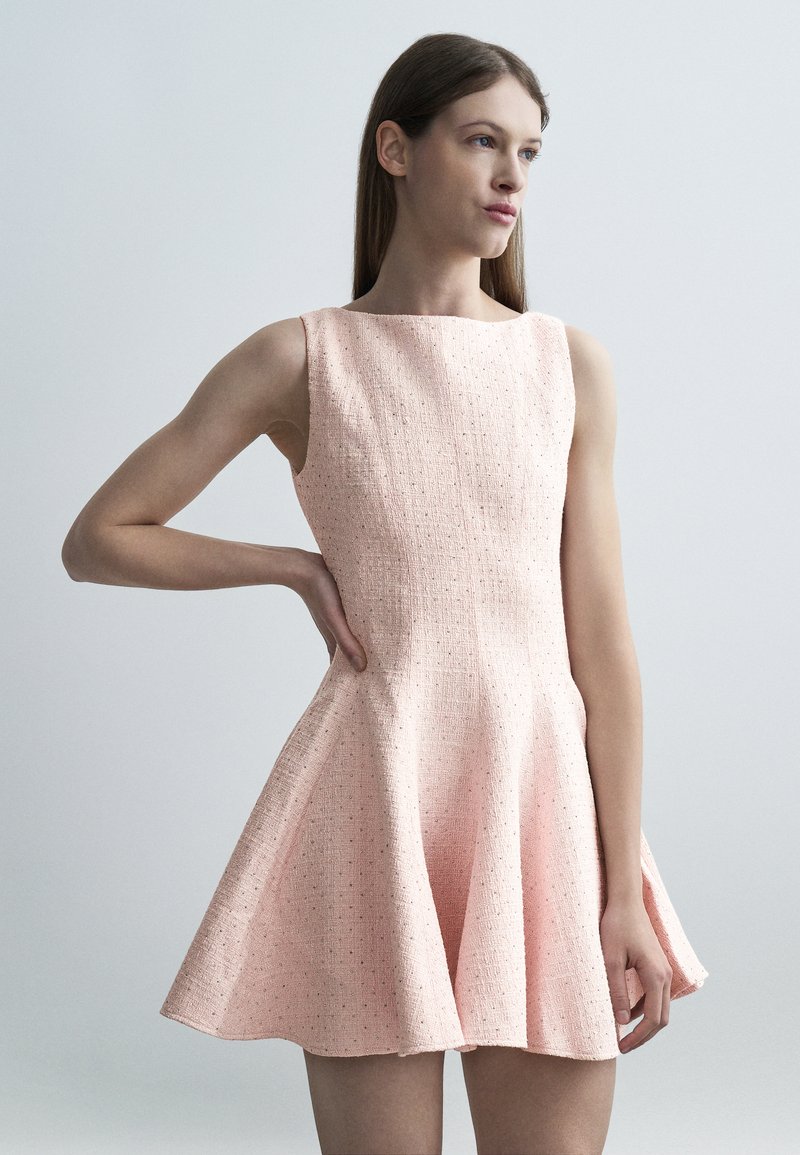 Γυναίκα που φορά ένα αμάνικο, ροζ φόρεμα με υφή και φουσκωτή φούστα, στέκεται με το ένα χέρι στη μέση της μπροστά σε απλό γκρι φόντο.