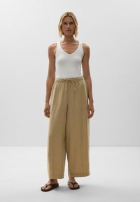 White ribbed tank top paired with loose, beige linen pants featuring an elastic waist and drawstring. Model wearing brown flip-flops.