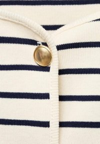 Cardigan lavorato a maglia con strisce verticali blu navy e bianche, con un primo piano di un bottone dorato e una texture a coste lungo il colletto e i bordi.