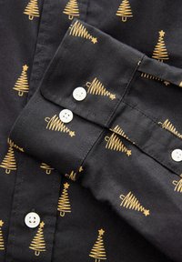 Chemise en coton noir avec un motif répétitif d'arbres de Noël dorés, dotée de boutons blancs aux poignets et au col pour la fermeture.