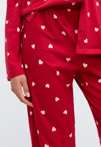 Κόκκινο σετ πυτζάμας με λευκό μοτίβο καρδιάς, φτιαγμένο από μαλακό ύφασμα. Διαθέτει ελαστι�κή μέση και οι άκρες των μανικιών έχουν αντίστοιχη λεπτομέρεια καρδιάς.