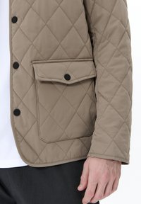 Chaqueta acolchada beige con un patrón de diamantes, botones a presión negros y un bolsillo frontal. Textura suave, con un ajuste entallado en el dobladillo.