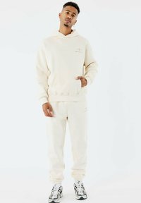 Junger Mann steht, trägt einen cremefarbenen Hoodie und passende Jogginghosen, weiße und schwarze Sneakers, vor einem schlichten weißen Hintergrund.