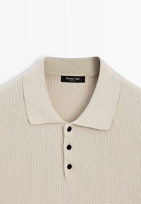 Polo en maille beige avec trois boutons noirs et un col côtelé, avec une étiquette Massimo Dutti à l’intérieur de l’encolure.