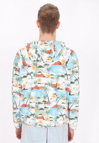 Letthæmmet hættejakke med et farverigt tropisk print med palmer, blomster og bjerge på en hvid baggrund.