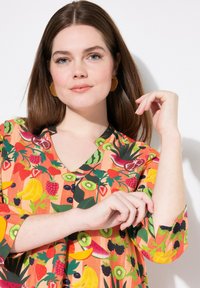 Blusa a fantasia di frutta colorata con scollatura a V e maniche a 3/4. Il tessuto appare liscio con uno sfondo arancione vivace e accenti verdi.