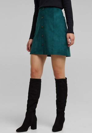 Femme portant une jupe boutonnée devant de couleur sarcelle, un haut noir à manches longues et des bottes noires à talons montant jusqu'aux genoux, sur un fond uni.
