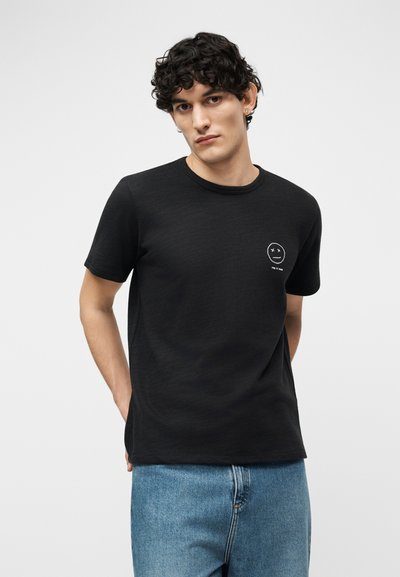 Άνδρας με σγουρά σκούρα μαλλιά, που φοράει μαύρο T-shirt με ένα μικρό λευκό γραφικό και ανοιχτόχρωμο τζιν, στέκεται με τα χέρια πίσω από την πλάτη.