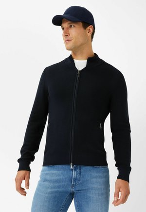 Navy ribbed zip-up jacket with a high collar, featuring side pockets and a fitted silhouette. Paired with light blue denim jeans.