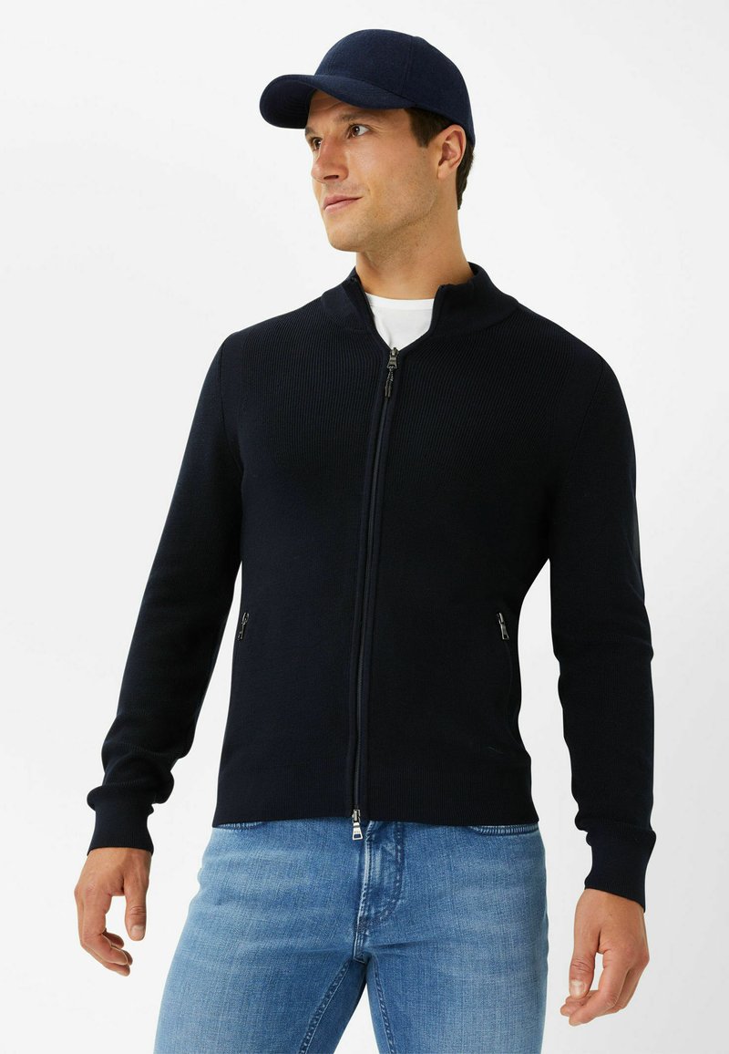Marineblau gerippte Zip-Jacke mit hohem Kragen, ausgestattet mit seitlichen Taschen und einer taillierten Silhouette. Kombiniert mit hellblauen Jeans.