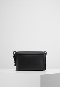 Černá obdélníková kožená kabelka přes rameno s klopou, nastavitelným ramenním popruhem a viditelným prošitím na bílém podstavci.