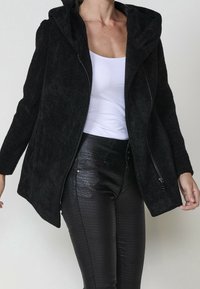 Femme portant une veste noire texturée zippée sur un débardeur blanc et un pantalon noir en cuir synthétique à motif reptile.