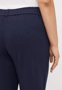 Granatowe spodnie z elastycznym pasem, gładką teksturą, dopasowanym krojem i boczną kieszenią; prezentowane od pasa w dół, widoczna ręka.