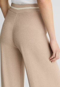 Pantaloni a gamba larga in tessuto a maglia beige chiaro. Presentano una vita a coste con una striscia bianca. Tessuto liscio con una vestibilità comoda.